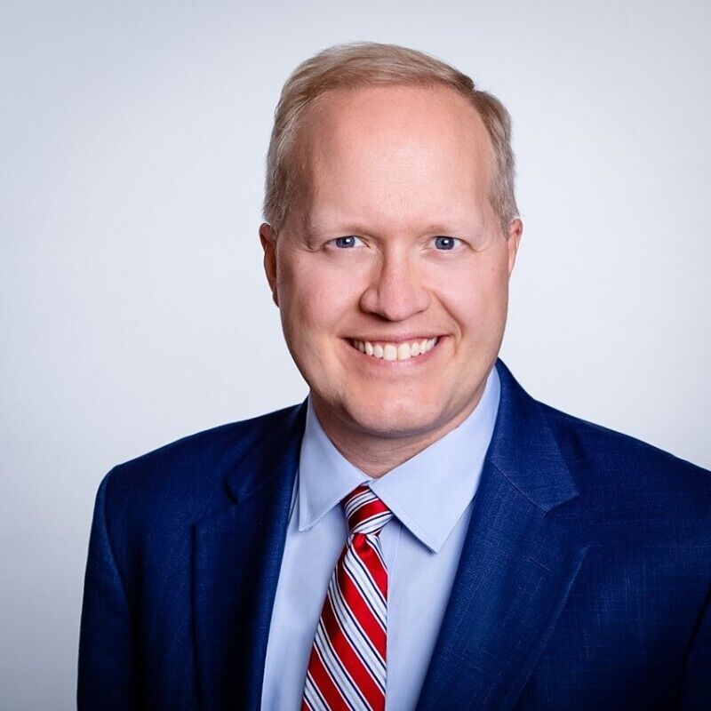 The image shows a man with fair skin and blond hair, wearing a blue suit jacket, a light blue dress shirt, and a red and blue striped tie. He is smiling and looking directly at the camera. The background is a plain white or light gray color. The man appears to be in his late 30s or early 40s.
